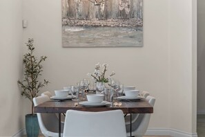 Una mesa de comedor dispuesta con platos, tazones y vasos blancos, rodeada de sillas blancas, con un cuadro encima y una planta a la izquierda.