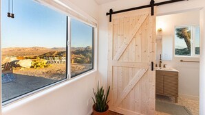 Una stanza con una porta a scorrimento in legno, un'ampia finestra che si affaccia su un paesaggio desertico, una pianta in vaso e un bagno con lavabo e specchio moderni.