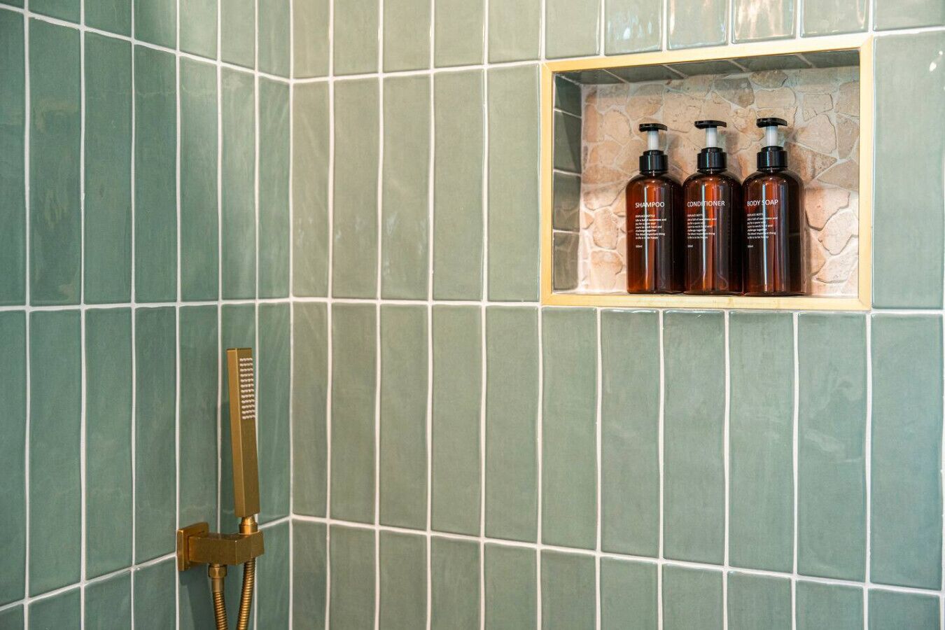 Een badkamer met groene tegels, een gouden douchekop en een plank met drie flessen, gelabeld als shampoo, conditioner en douchezeep.