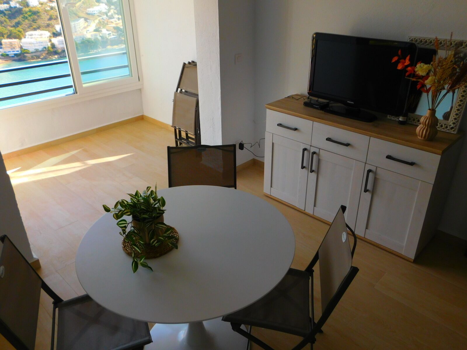Un coin repas avec une table ronde, des chaises, une plante en pot, un téléviseur posé sur un meuble en bois et une vue sur la mer par la fenêtre.