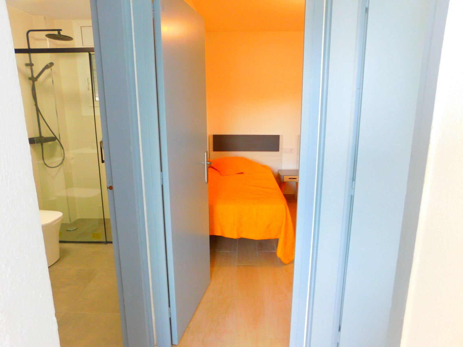 A narrow hallway leading to a room with a bed, a bedside table, and a shower area.