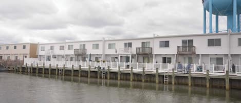 Fila de edificios blancos junto al agua con balcones, un muelle y una torre de agua azul al fondo.
