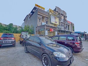 A parking lot with several cars, including a black sedan and a maroon SUV. In the background, there are multi-story buildings with various signs, including one for a restaurant and another for a bank. - OYO 90615 Jojo Homestay (Padang)