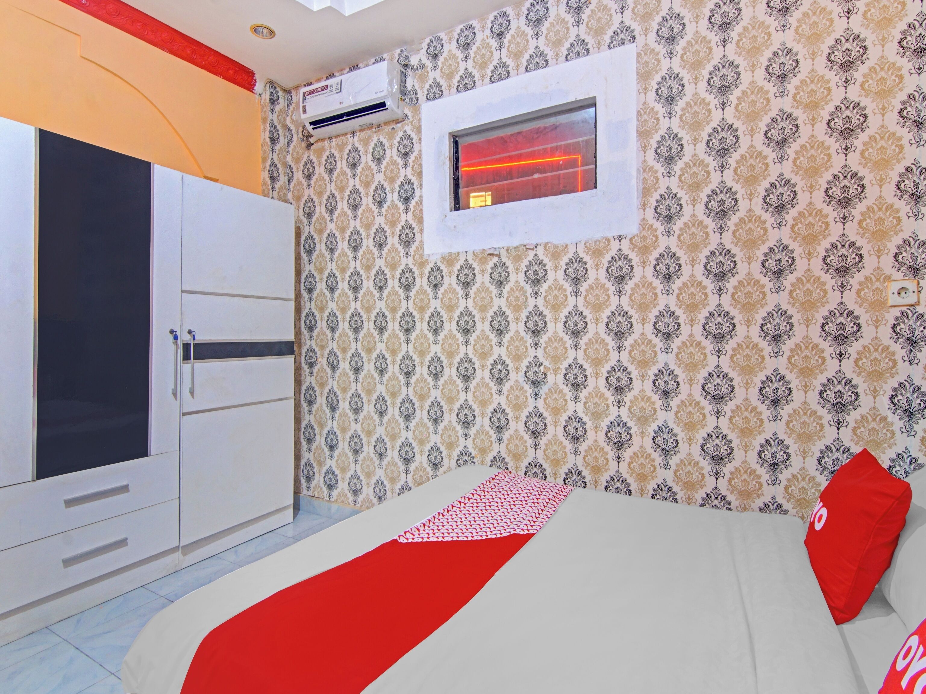 A hotel room with a wardrobe, a bed with a red pillow, a window with a red neon light, and patterned wallpaper.