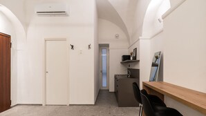 Un pasillo con una puerta blanca, una kitchenette pequeña con un lavamanos y un microondas, y un mostrador de madera con dos sillas negras.