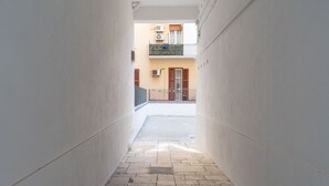 Un pasillo estrecho con suelo de baldosas que conduce a un edificio con balcón y unidad de aire acondicionado.