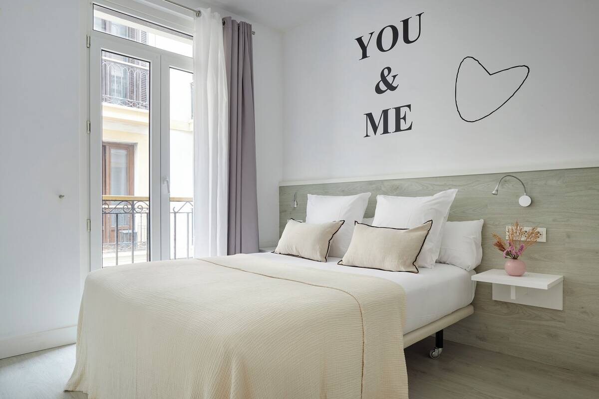 A bedroom with a bed, a wall with "YOU & ME" and a heart, a nightstand with a lamp, and a window with curtains.