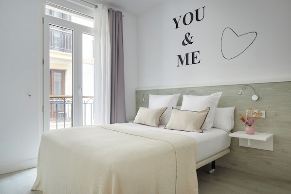 Un dormitorio con una cama, una pared con la inscripción “YOU & ME” y un corazón, una mesita de noche con una lámpara, y una ventana con cortinas.