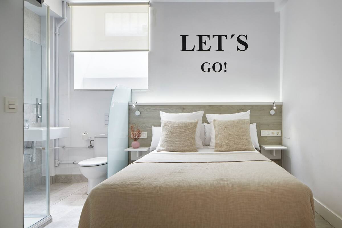 A modern bedroom with a bed, a nightstand, a bedside table, a window, and a bathroom visible in the background.