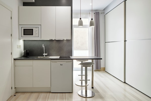 A compact kitchen with a built-in microwave, a coffee maker, and a small refrigerator. There is a bar stool and a window with curtains.