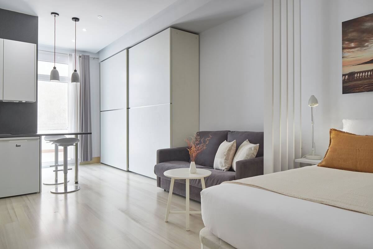 A modern hotel room with a bed, a sofa, a small table, and a kitchenette area.