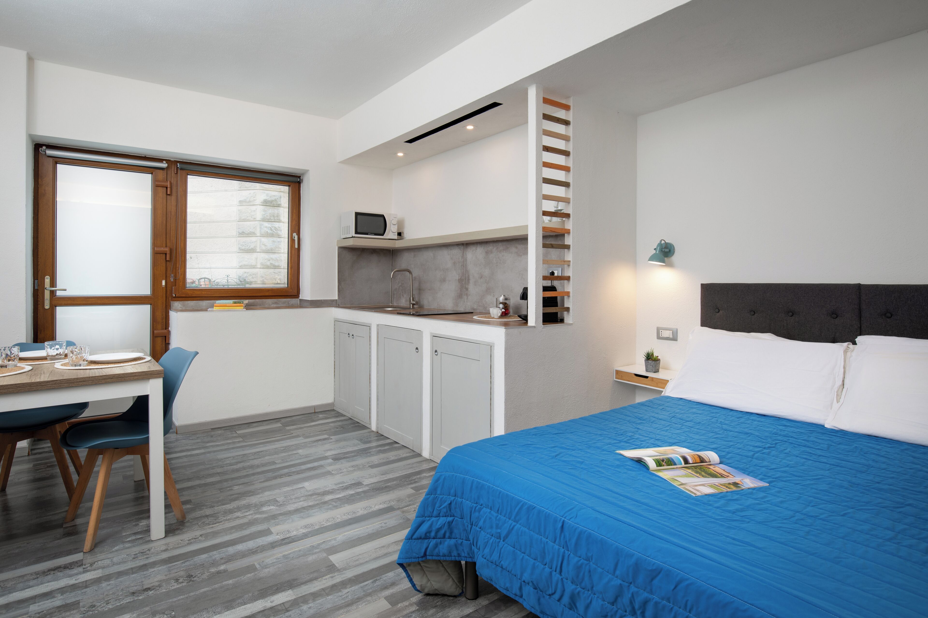 A compact room with a bed, a desk with a chair, a kitchenette, and a window.
