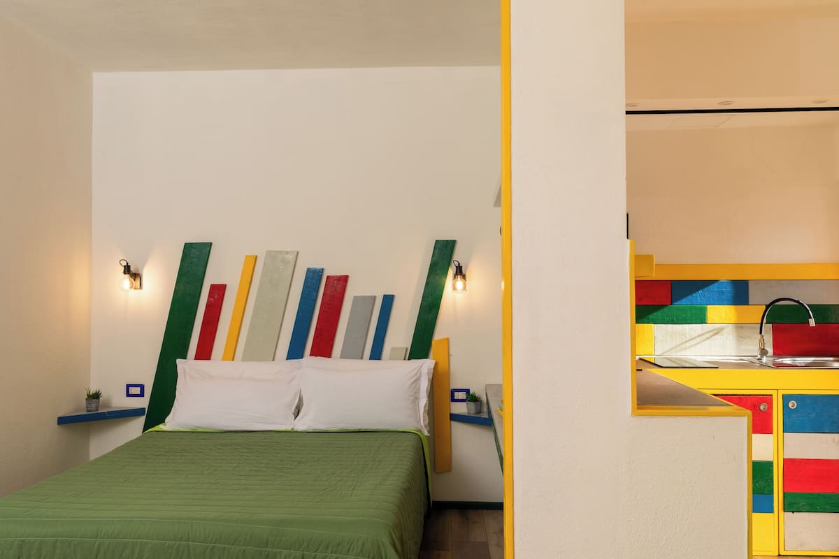 Ein Zimmer mit einem Bett, einem farbenfrohen Wandbild, einem kleinen Regal mit einer Pflanze und einer Küchenzeile mit Spüle und Schränken.