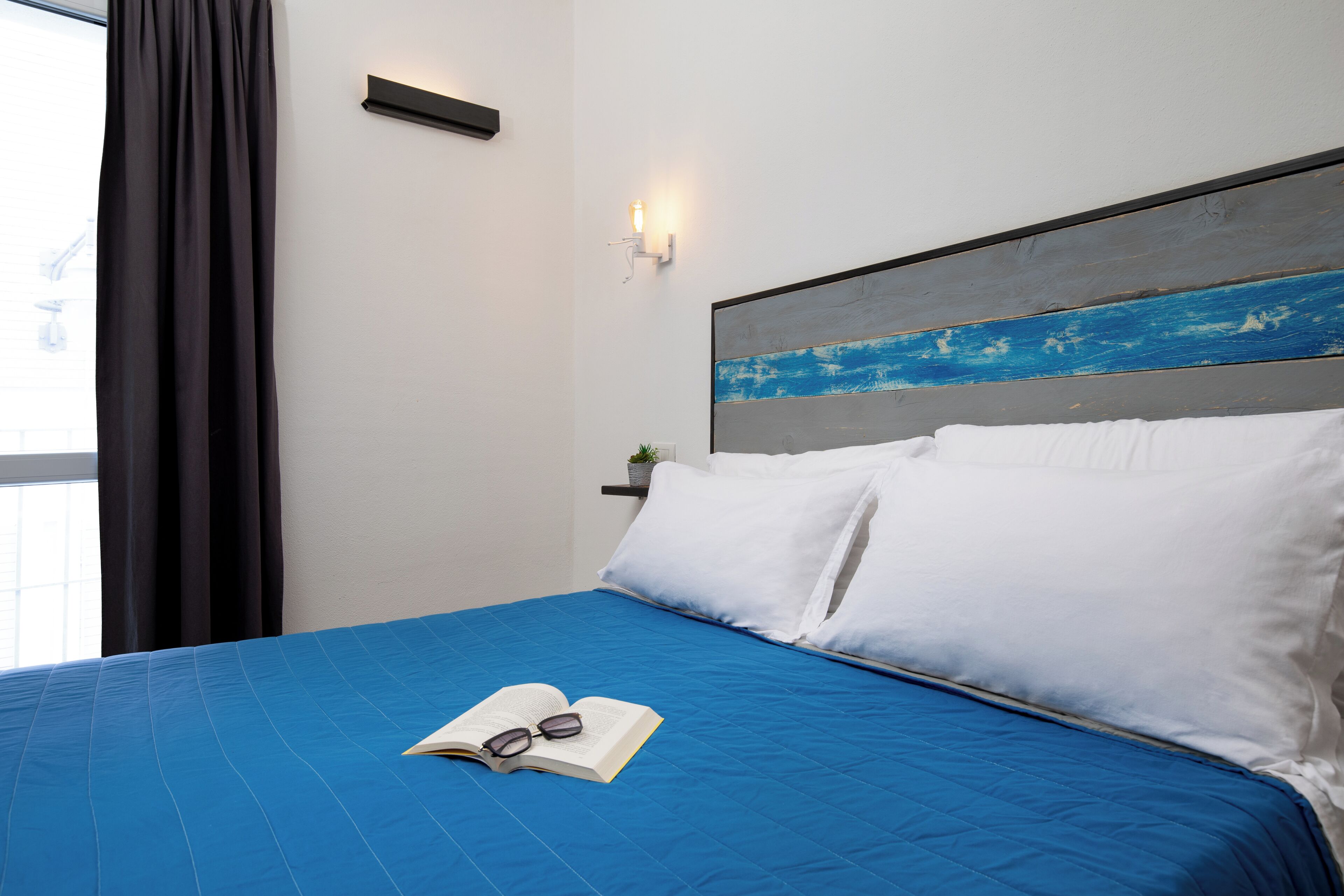A neatly made bed with a blue bedspread, white pillows, and a book with glasses on it.