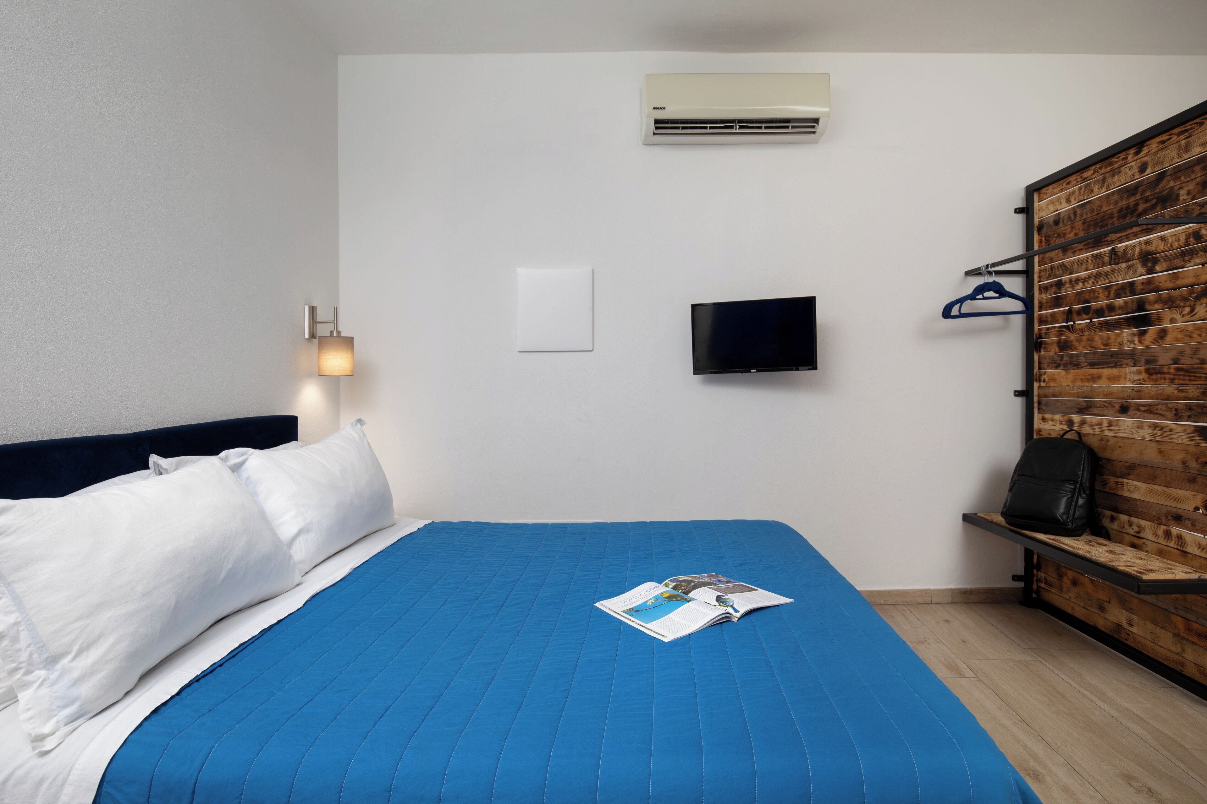 A hotel room with a bed, a flat-screen TV, an air conditioner, a wooden bench, and a wall-mounted light fixture.