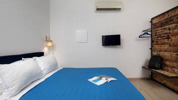 A hotel room with a bed, a flat-screen TV, an air conditioner, a wooden bench, and a wall-mounted light fixture.