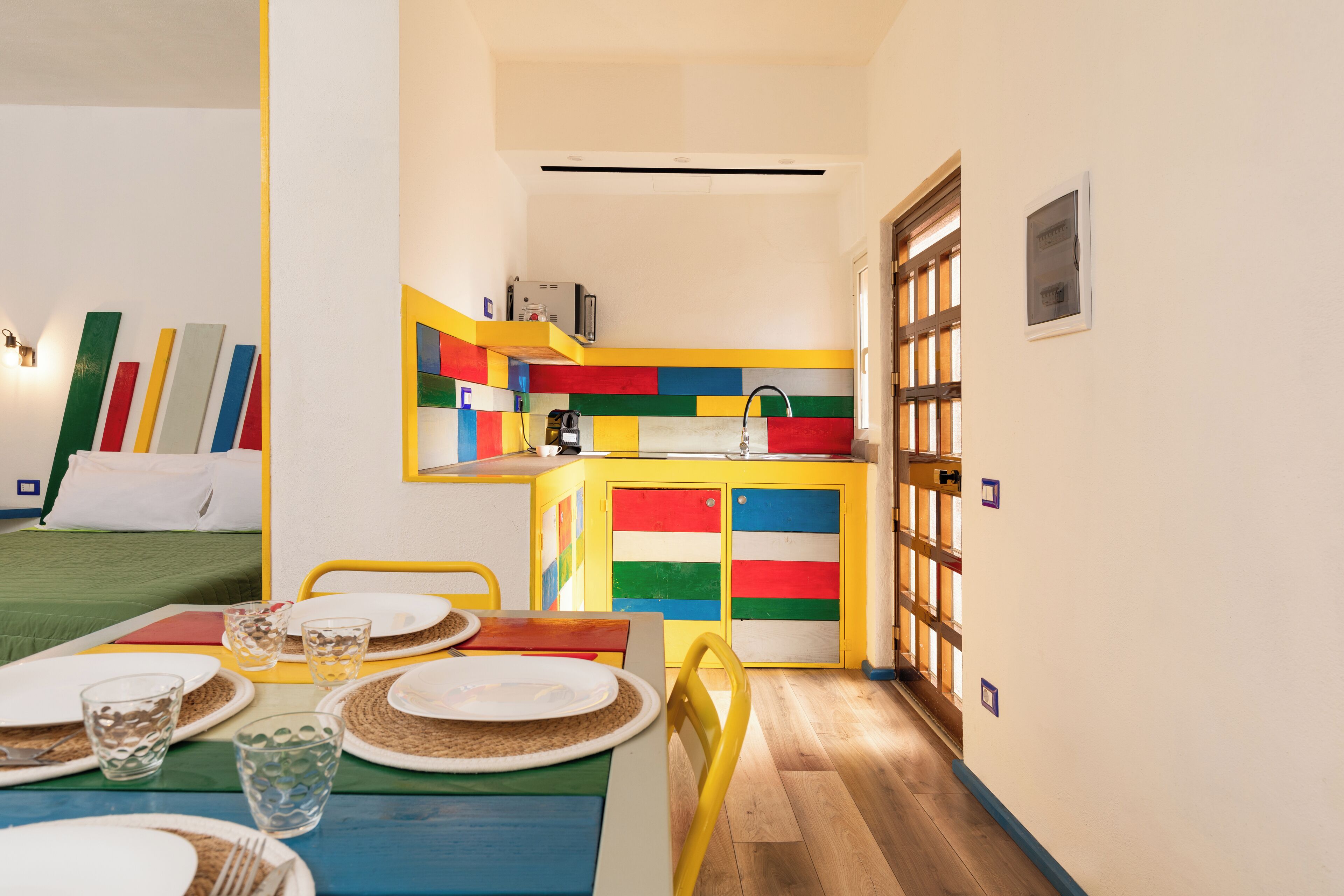 A brightly colored room with a dining table set for two, a kitchenette with colorful cabinets, and a bed with green bedding.