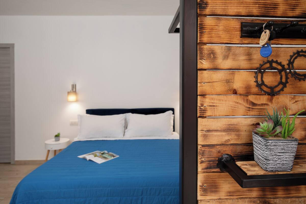 Ein Schlafzimmer mit blauer Bettdecke, einem kleinen Nachttisch mit Lampe und einem an der Wand befestigten Regal mit Pflanze.