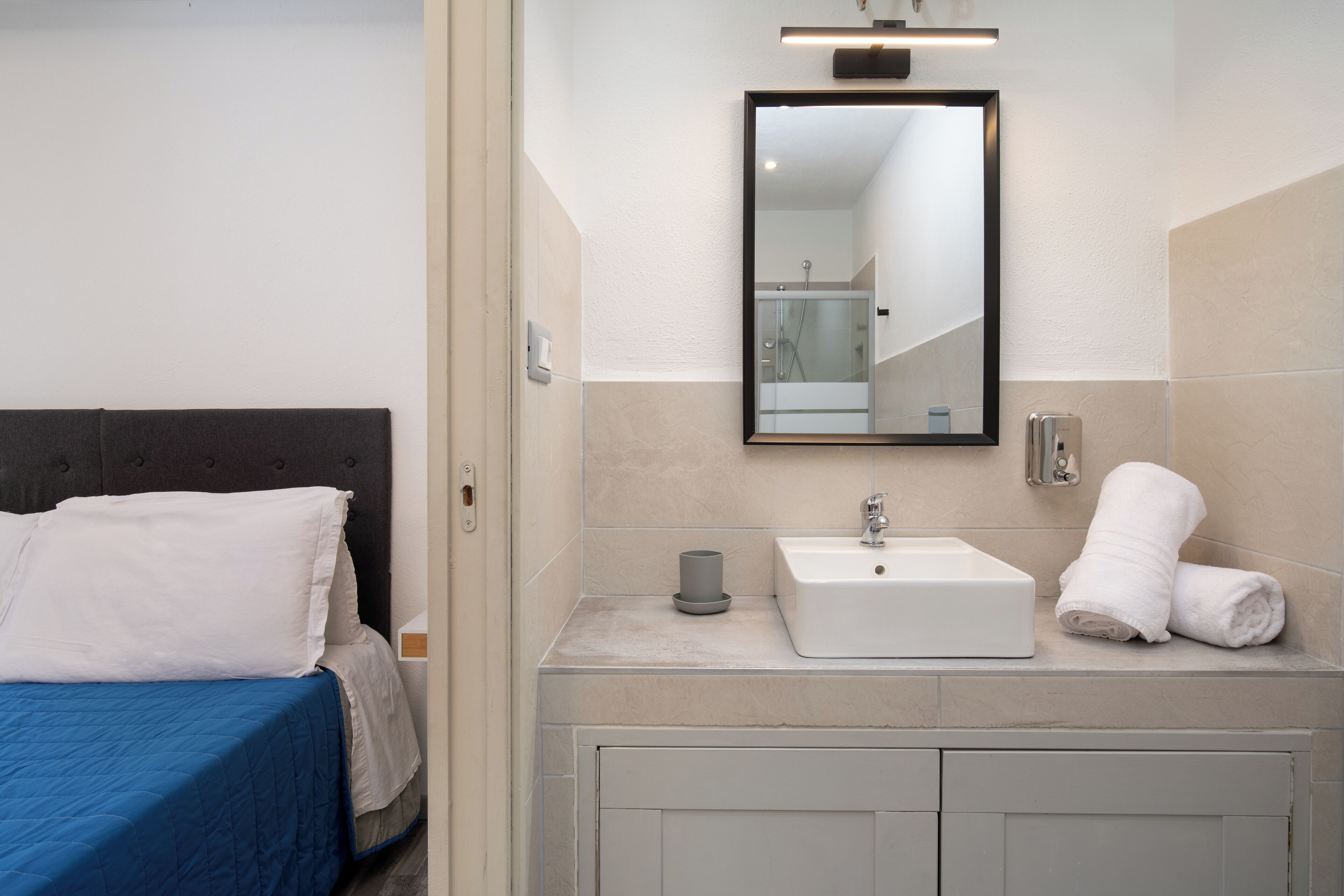Una camera d'albergo moderna con un letto, un bagno con lavandino e specchio, e un asciugamano sul ripiano.