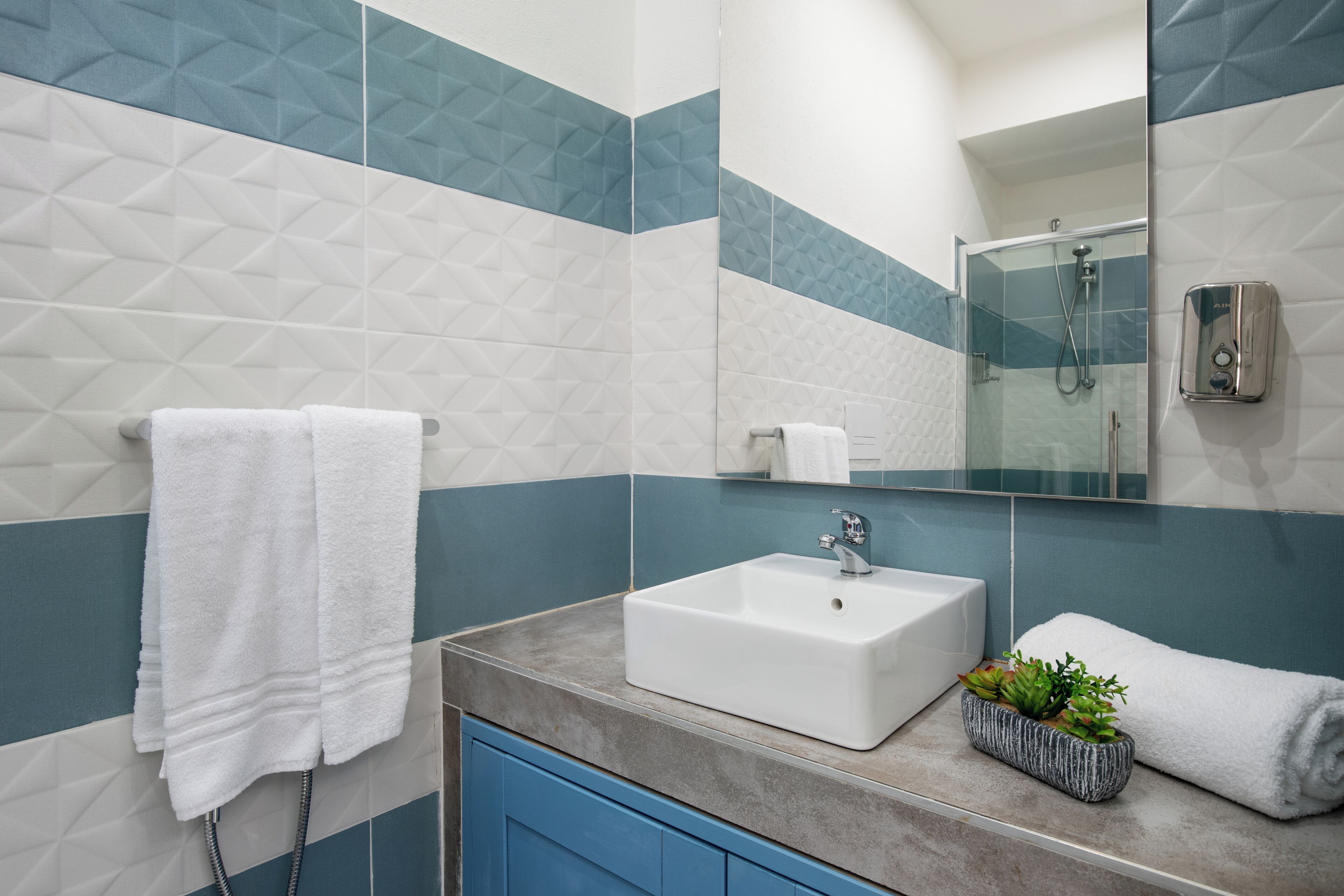 A bathroom with a white sink, blue and white tiled walls, a glass shower enclosure, and a small potted plant on the countertop.