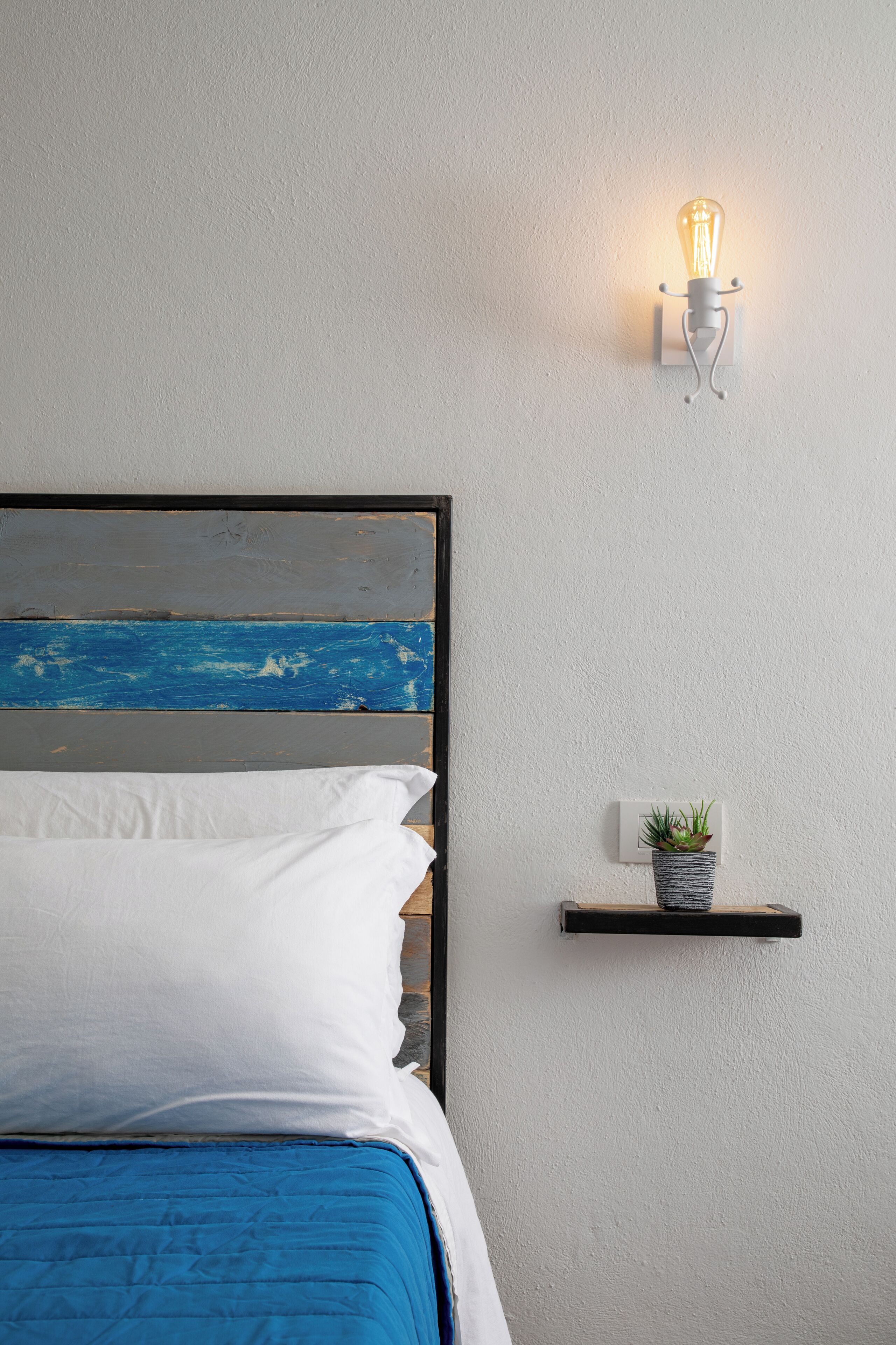 A bed with a blue bedspread, white pillows, and a wooden headboard with a painted ocean view. A wall-mounted light fixture and a small shelf with a plant are also visible.