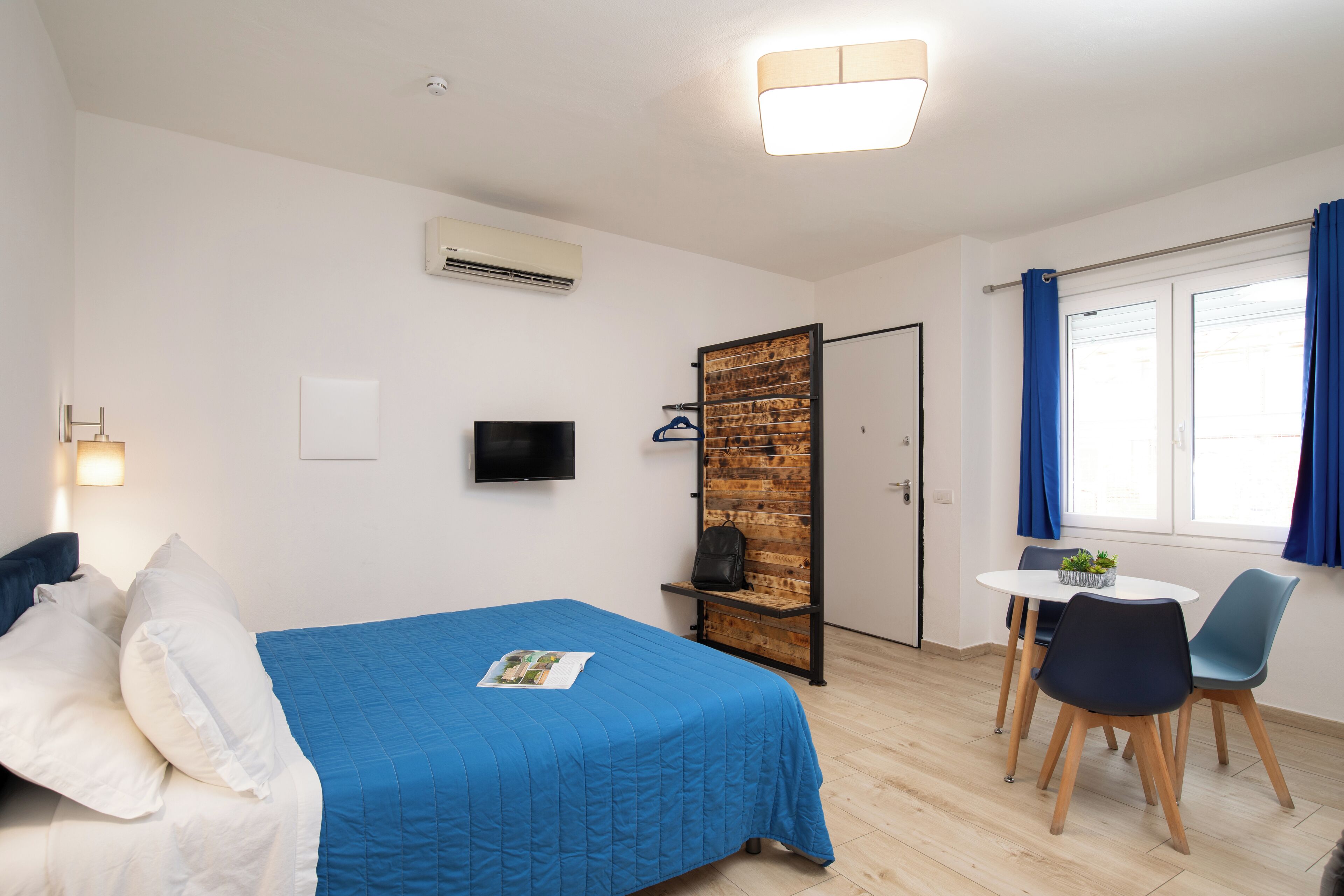 A bedroom with a bed, a TV, a wooden wall, a door, and a dining area with chairs and a table.