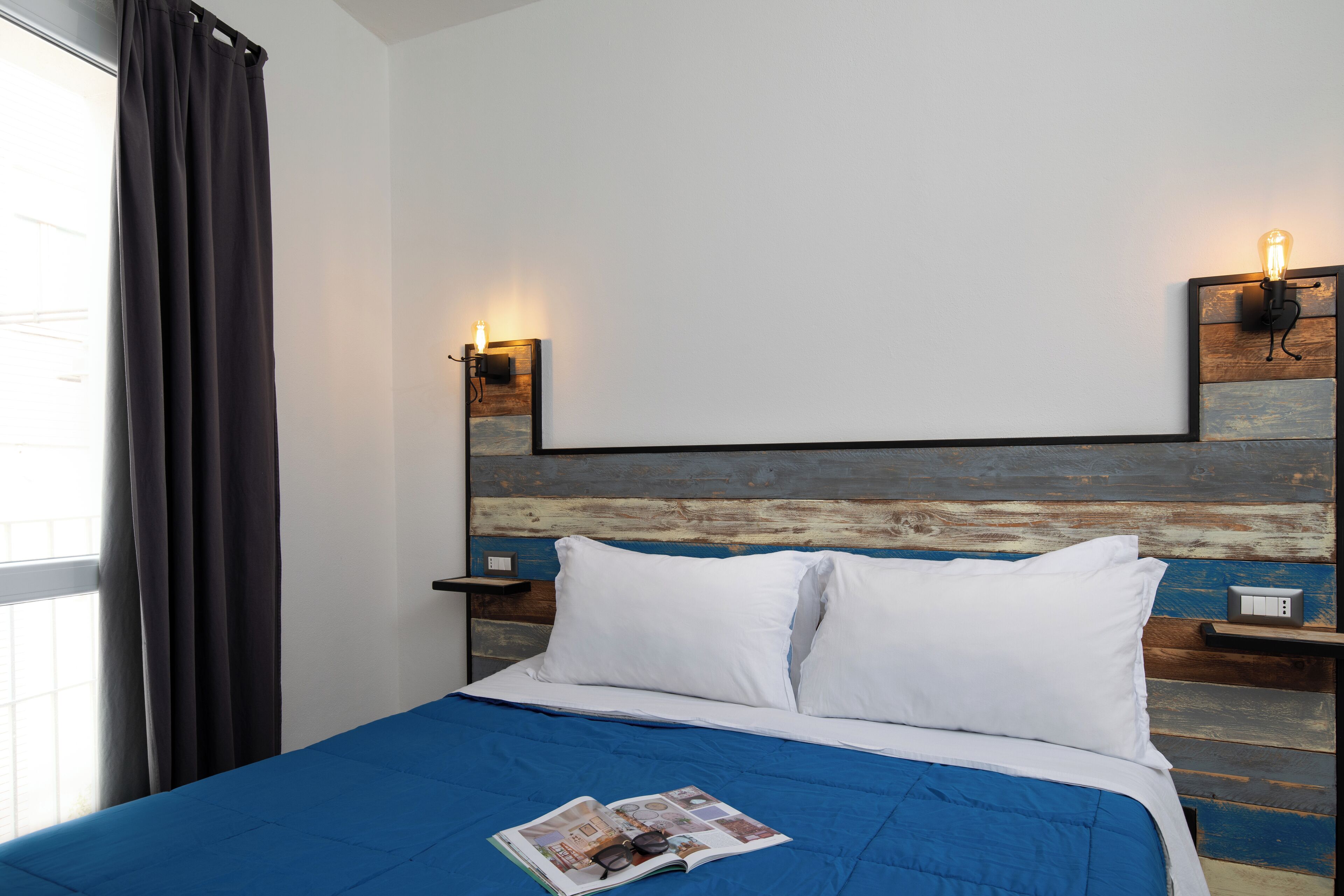 Ein ordentlich gemachtes Bett mit blauer Bettdecke, weißen Kissen und einer Zeitschrift darauf. Zwei Nachttische mit Lampen stehen bereit, darüber ein Wandleuchter.