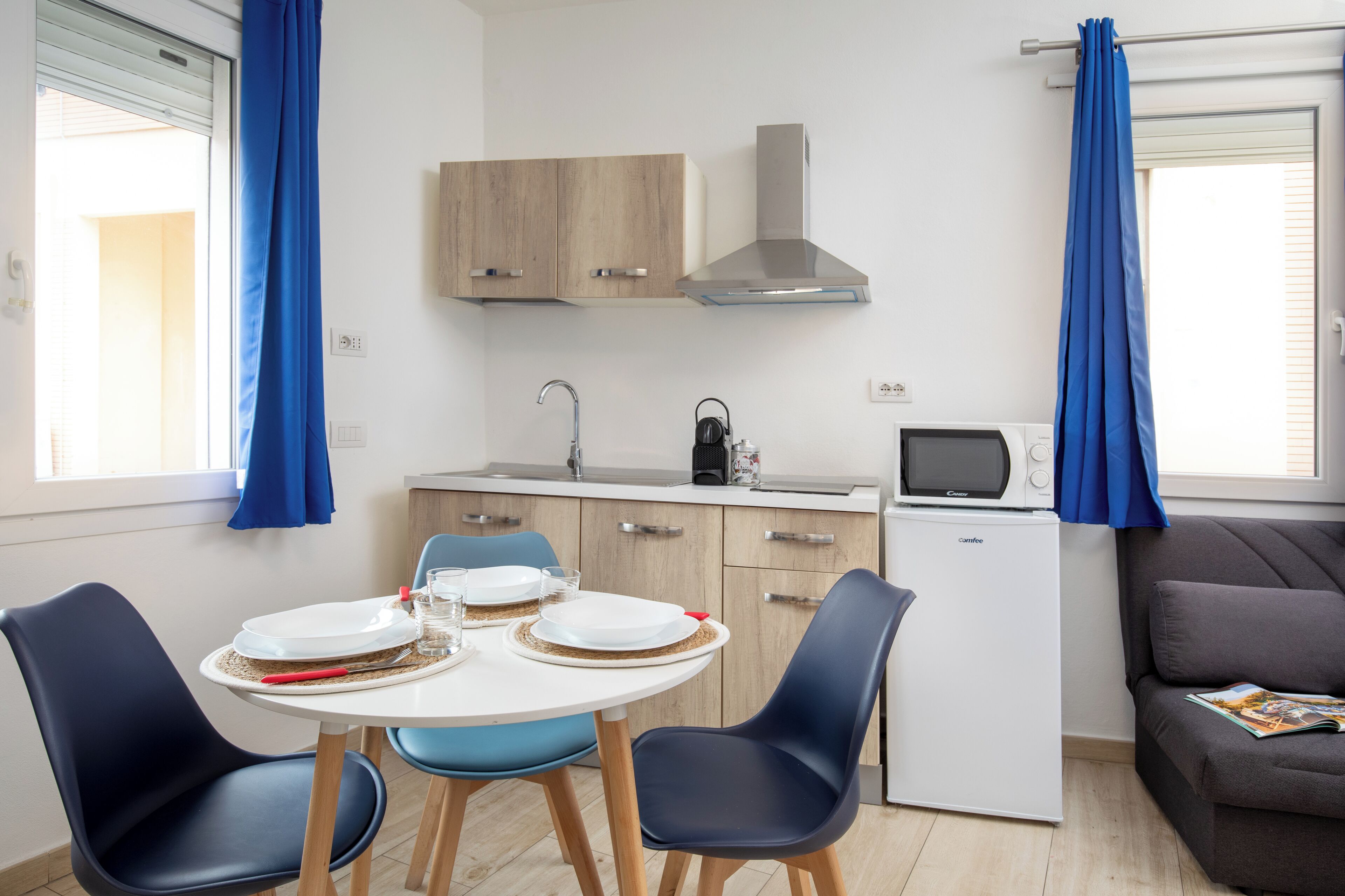 A compact kitchen with a microwave, kettle, and a small refrigerator. There is a dining table with plates and glasses, and a sofa in the living area.
