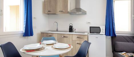 A compact kitchen with a microwave, kettle, and a small refrigerator. There is a dining table with plates and glasses, and a sofa in the living area.