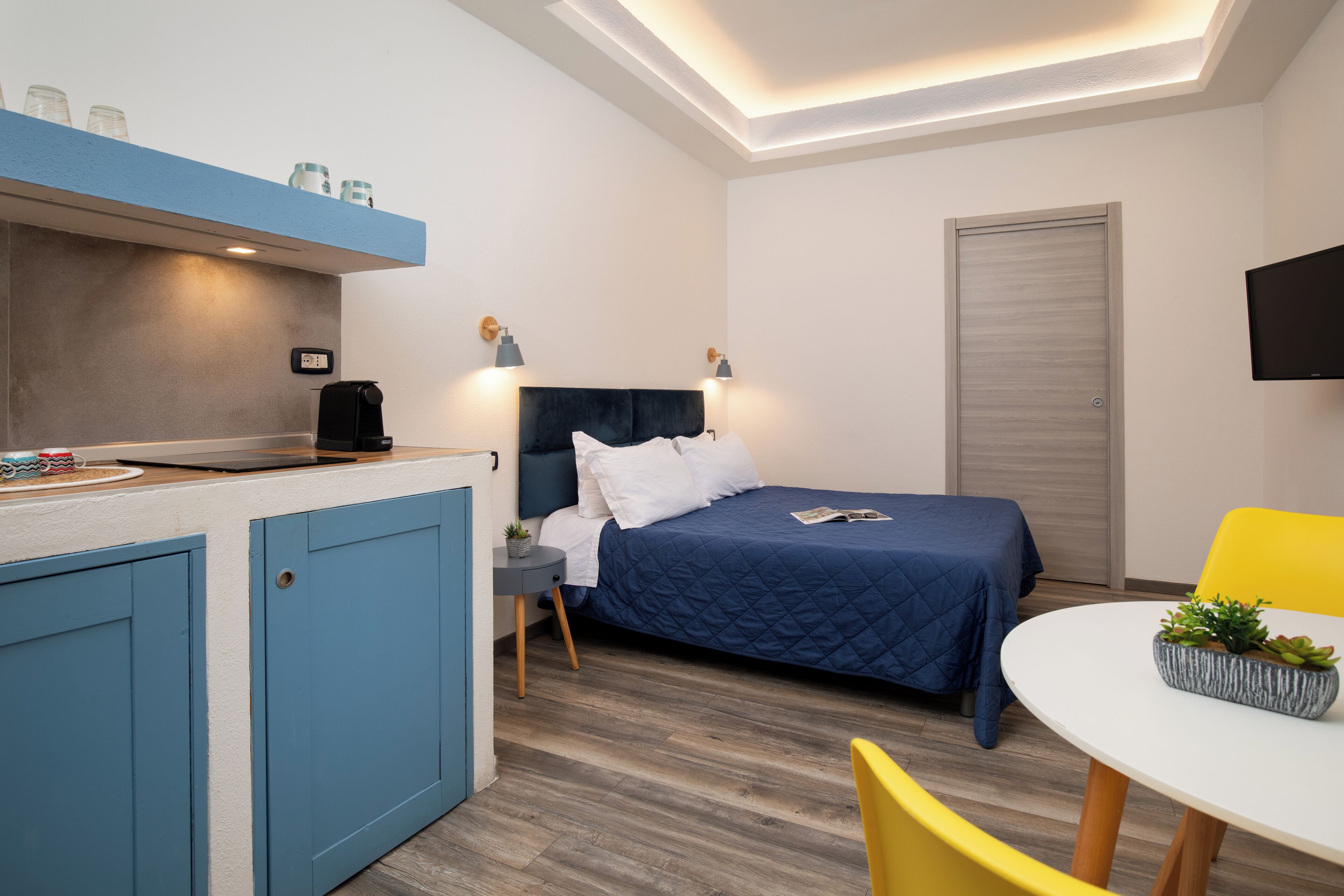 Une pièce de taille modeste comprenant un lit, une petite table, une chaise, un coin cuisine et un téléviseur fixé au mur.