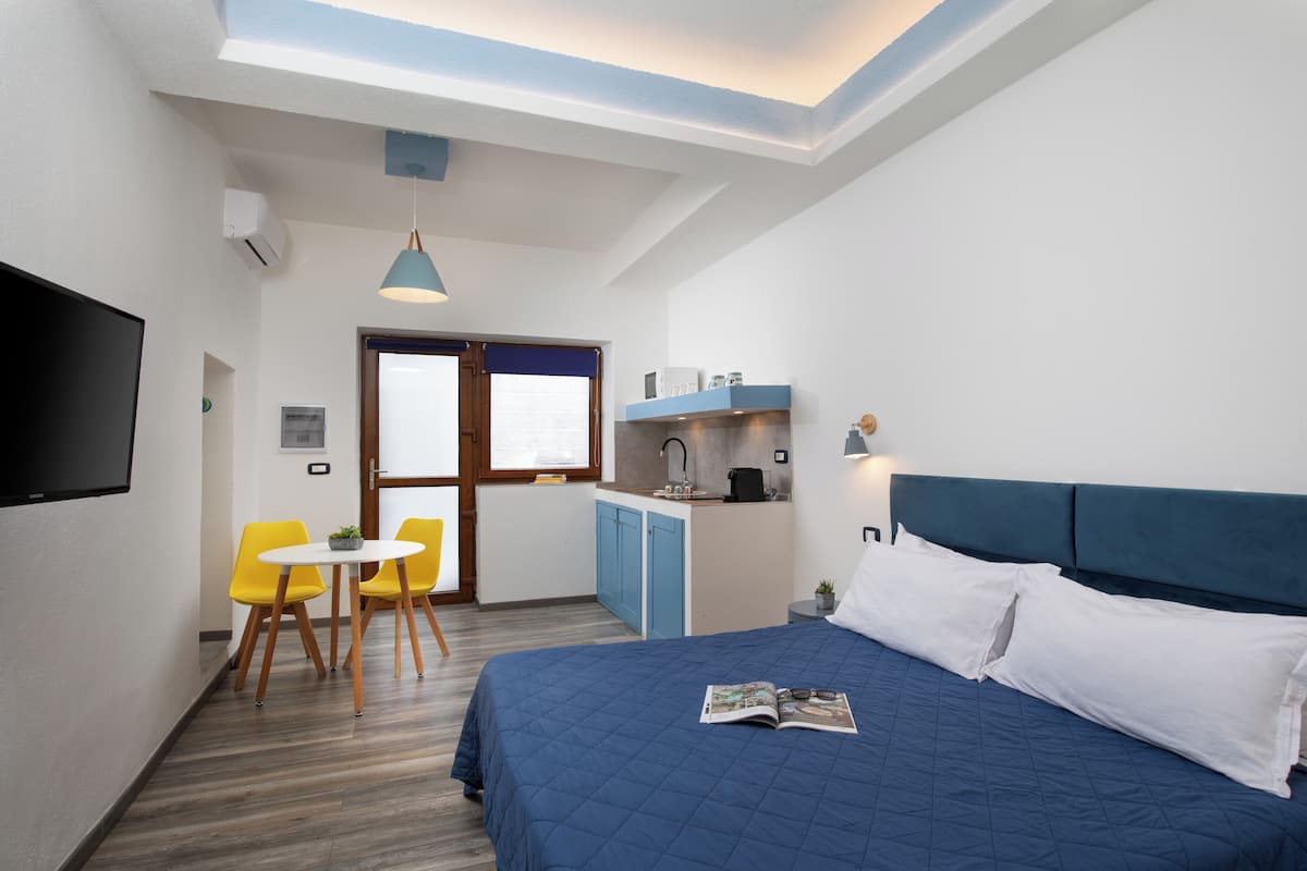 Ein Hotelzimmer mit einem Bett, einem Schreibtisch mit Stuhl, einer kleinen Küchenzeile und einem an der Wand befestigten Fernseher.