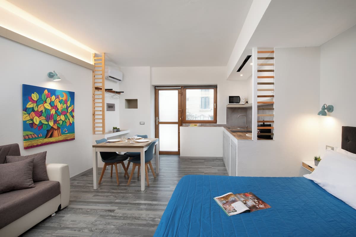 Ein modernes Schlafzimmer mit blauer Bettdecke, einem Essbereich mit Tisch und Stühlen, einem farbenfrohen Bild an der Wand und einer Treppe, die zu einer oberen Ebene führt.