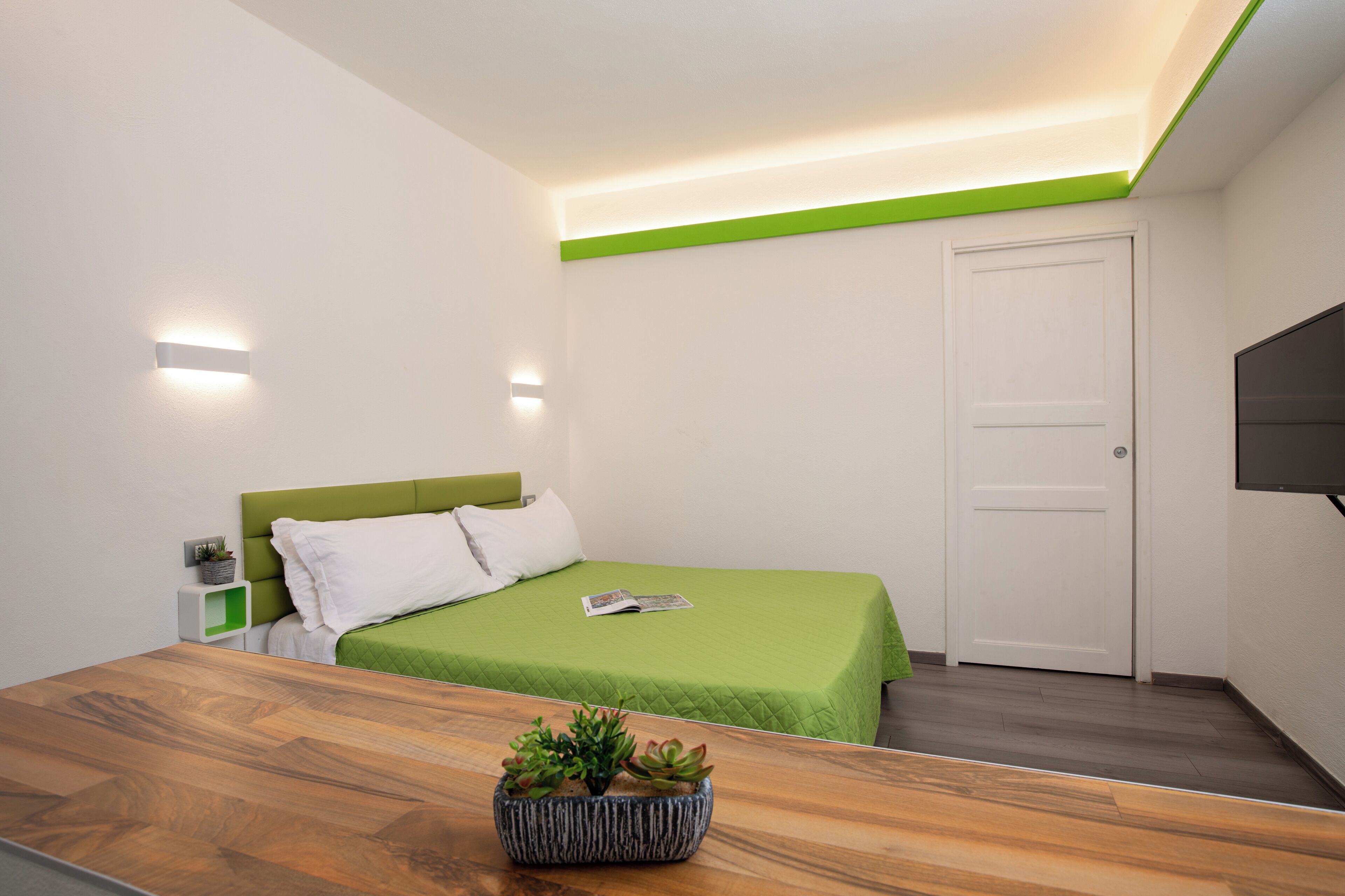 Ein modernes Schlafzimmer mit einer grünen Bettdecke, einem Nachttisch aus Holz mit einer Pflanze, einer weißen Tür und einem an der Wand befestigten Flachbildfernseher.