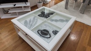 Une table basse avec un plateau en verre et un socle blanc, entourée d’un tapis blanc et posée sur un parquet.