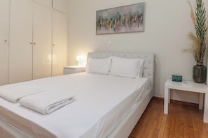 A neatly made bed with white linens and a folded towel, a bedside table with a lamp, and a wall-mounted abstract painting. - Charming 1 Bd Apt near ASOE in Athens (Athens)
