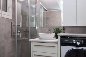 A modern bathroom with a glass-enclosed shower, a white sink, and a washing machine. - Charming 1 Bd Apt near ASOE in Athens (Athens)