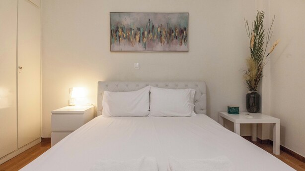 Une chambre à coucher bien rangée, avec un grand lit, une table de chevet avec une lampe, une petite table avec un vase et un tableau accroché au mur.