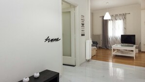 Un salon moderne avec un mur blanc, un banc noir, un téléviseur posé sur un meuble et une porte donnant sur une autre pièce.