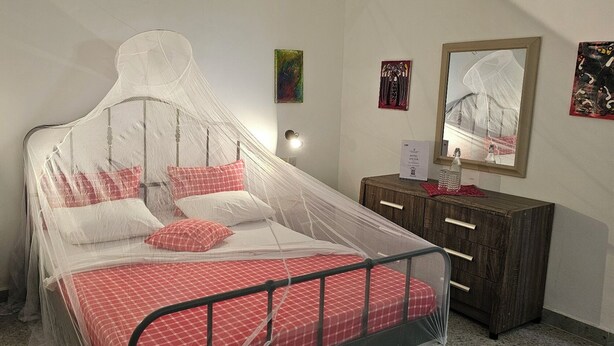 A bed with a mosquito net, a wooden nightstand, a mirror, and a small cabinet with drawers.