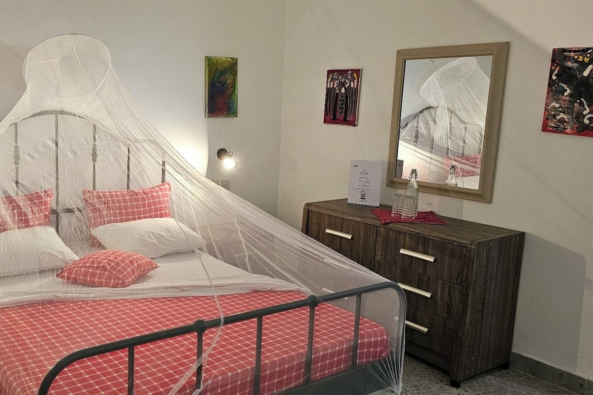 Uma cama com mosquiteiro, mesa de cabeceira de madeira, espelho e um pequeno armário com gavetas.