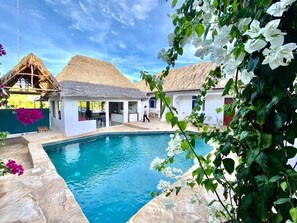 A swimming pool with a stone border, surrounded by white buildings with thatched roofs, and lush greenery. - Dragonfly (Jambiani)