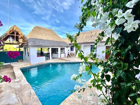 A swimming pool with a stone border, surrounded by white buildings with thatched roofs, and lush greenery.