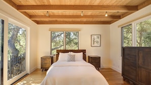 Une chambre avec un plafond en bois, de grandes fenêtres, un lit avec du linge de lit blanc, une table de chevet et une commode.