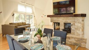 Un salon moderne avec une table à manger en verre pour quatre personnes, une cheminée en pierre, un canapé confortable et un grand téléviseur à écran plat fixé au-dessus de la cheminée.
