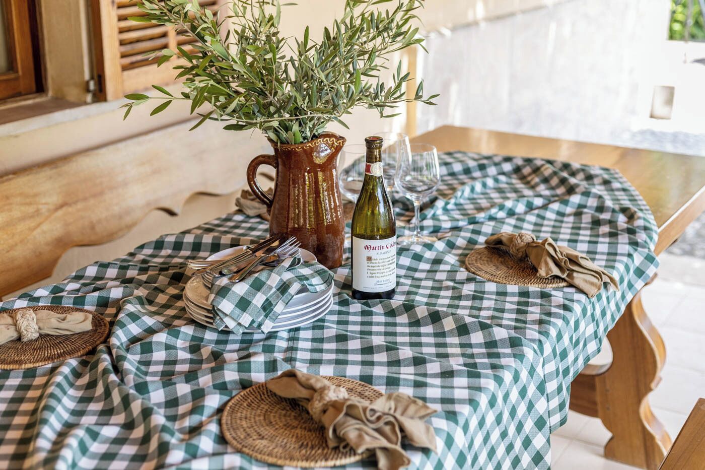 Una mesa dispuesta con mantel a cuadros, una botella de vino, un florero con follaje y diferentes utensilios de mesa.