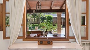 Une fenêtre avec des cadres en bois et des rideaux blancs, donnant sur un jardin où l’on peut voir des plantes en pot et une table avec des verres à vin et des bouteilles.