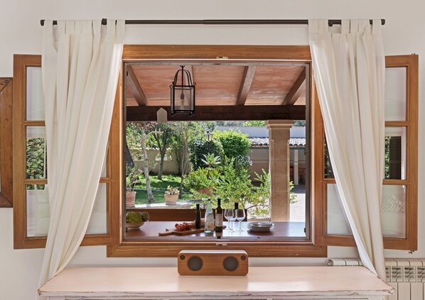 A window with wooden frames and white curtains, overlooking a garden with potted plants and a table with wine glasses and bottles.