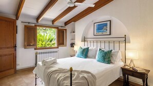 Une chambre à coucher avec un lit, une fenêtre avec des volets, un ventilateur de plafond, un radiateur et une table de chevet avec une lampe.