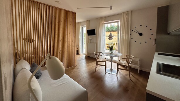 A modern living room with a sofa, a dining table with chairs, a kitchen area, and a large window with curtains. - Charmantes Design-appartement für 2 Personen in Utting am Ammersee (Utting am Ammersee)