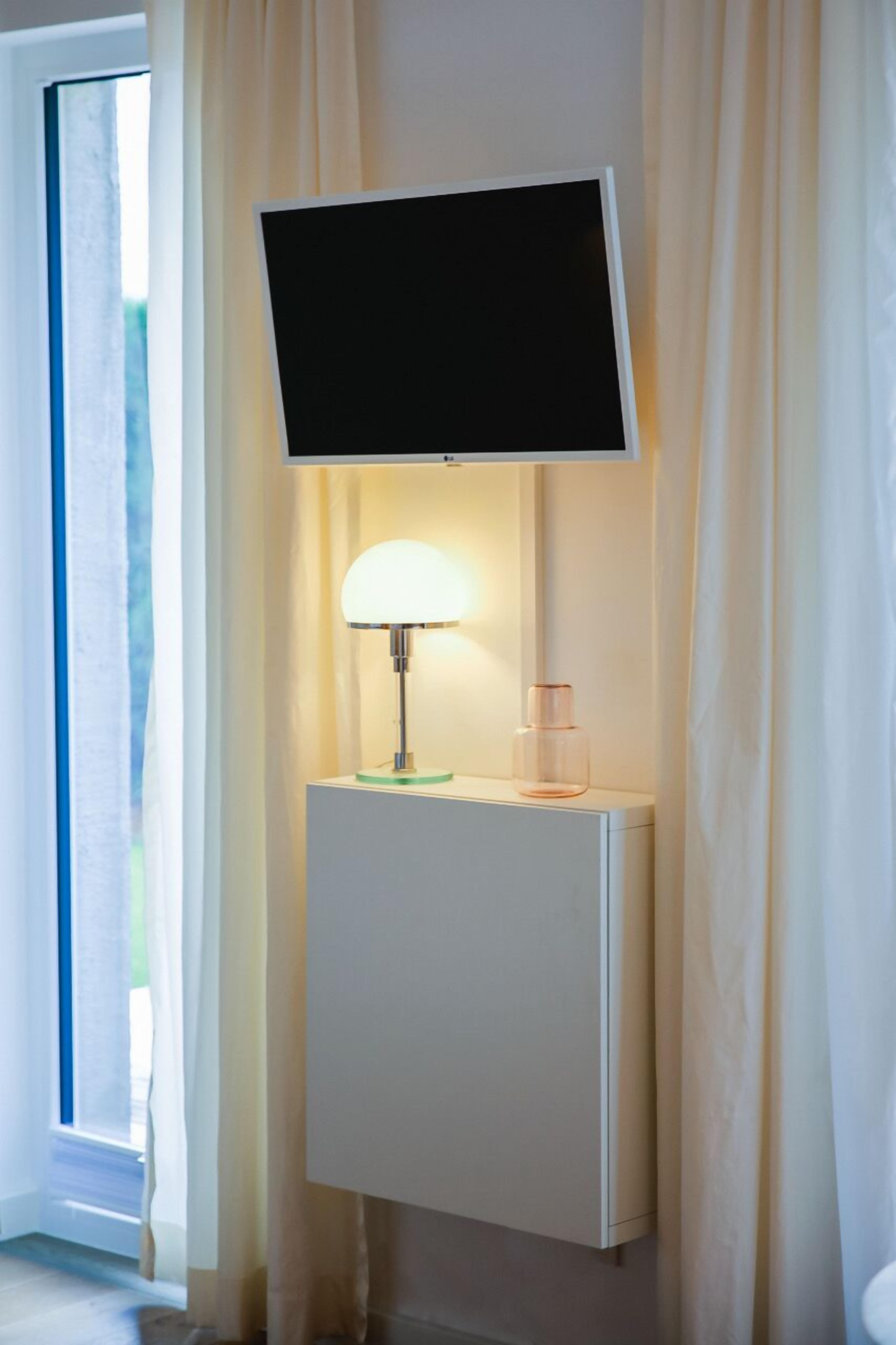Un téléviseur fixé au mur, au-dessus d’un meuble blanc, avec une lampe et une bouteille en verre.